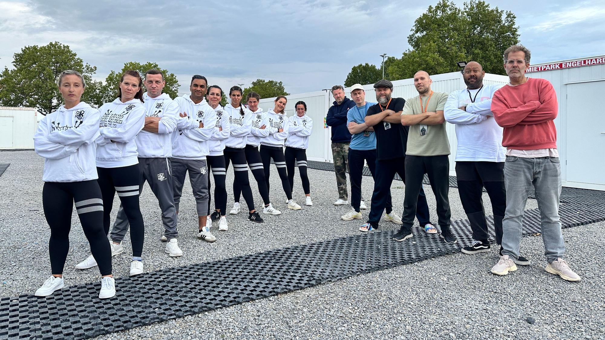 Das Bild zeigt die jungen Sportler aus Brühl, die mit verschränkten Armen in einer Reihe posieren. Daneben bilden die Musiker der Gruppe Deichkind eine Reihe.