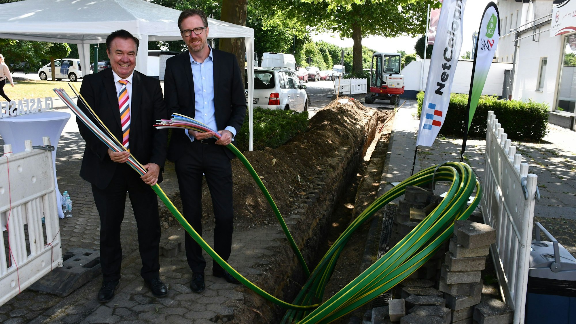 Zwei Männer im Anzug halten dicke Bündel von Glasfasern vor einer Baugrube in den Händen.