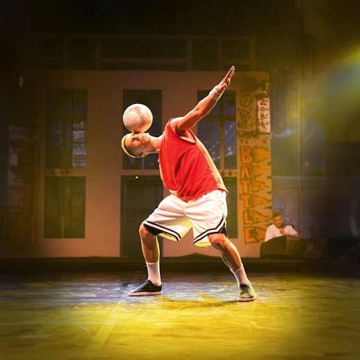 Das Bild zeigt den Künstler in kurzer Sportbekleidung auf der Bühne, wie er einen Fußball auf dem Kopf balanciert.