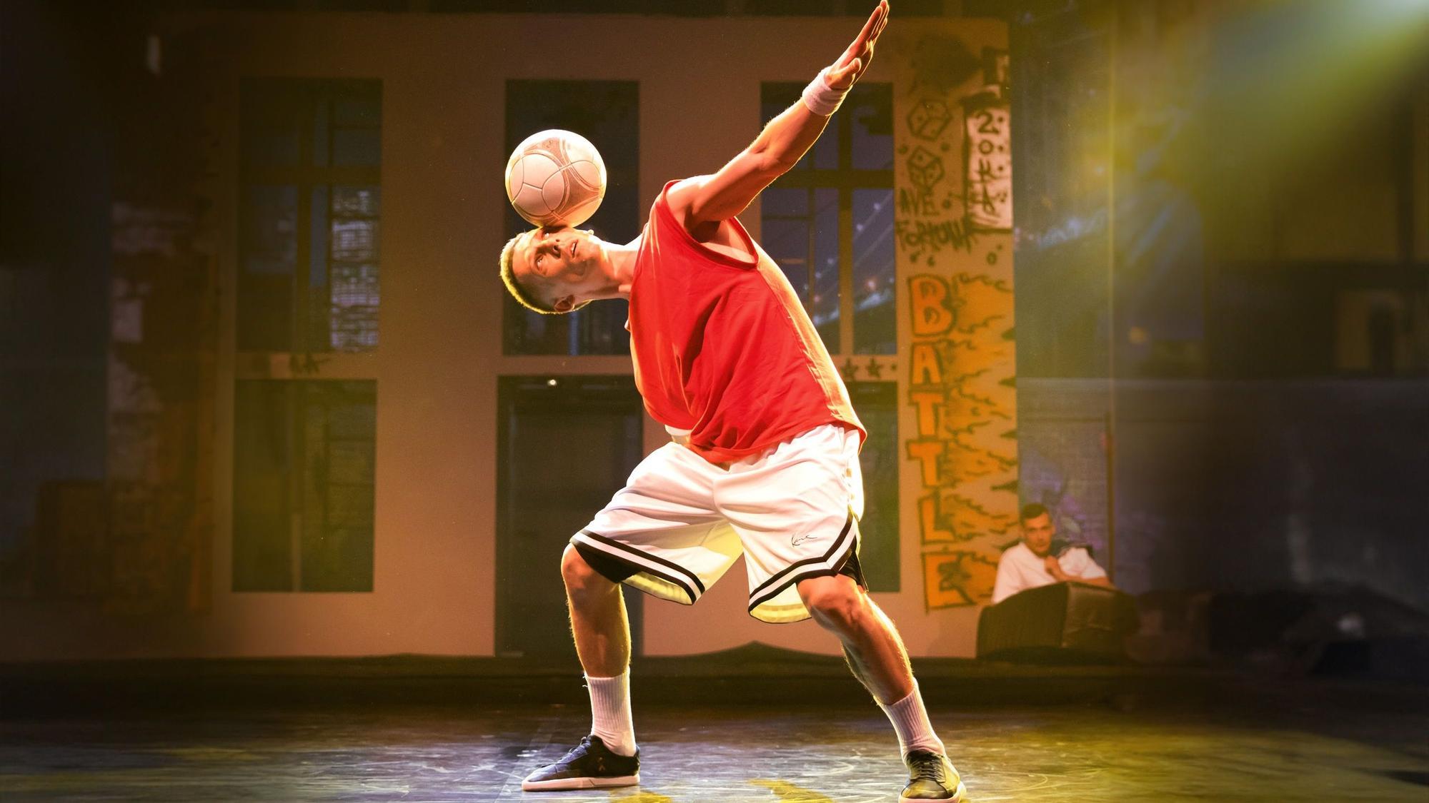 Das Bild zeigt den Künstler in kurzer Sportbekleidung auf der Bühne, wie er einen Fußball auf dem Kopf balanciert.