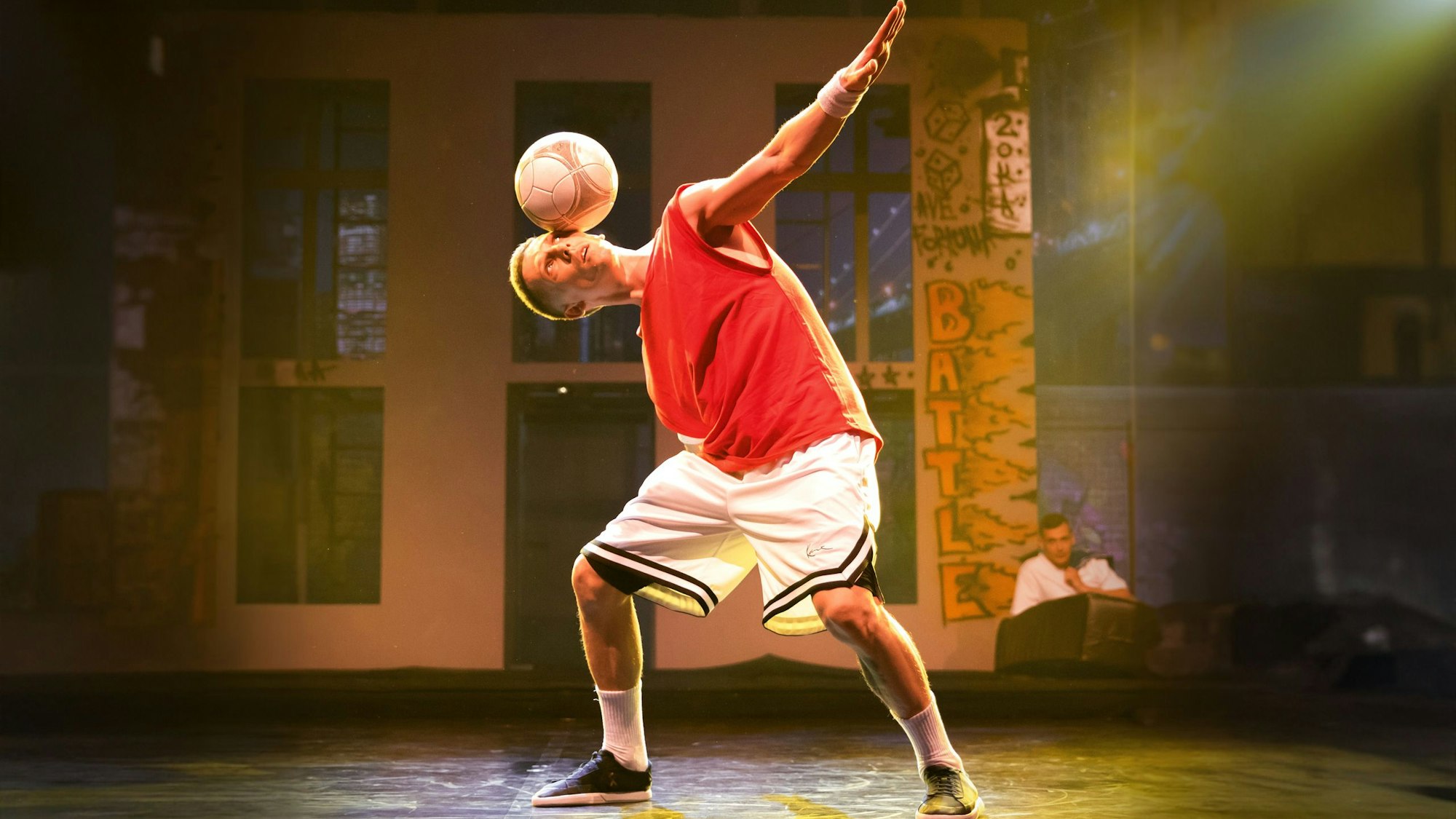 Das Bild zeigt den Künstler in kurzer Sportbekleidung auf der Bühne, wie er einen Fußball auf dem Kopf balanciert.