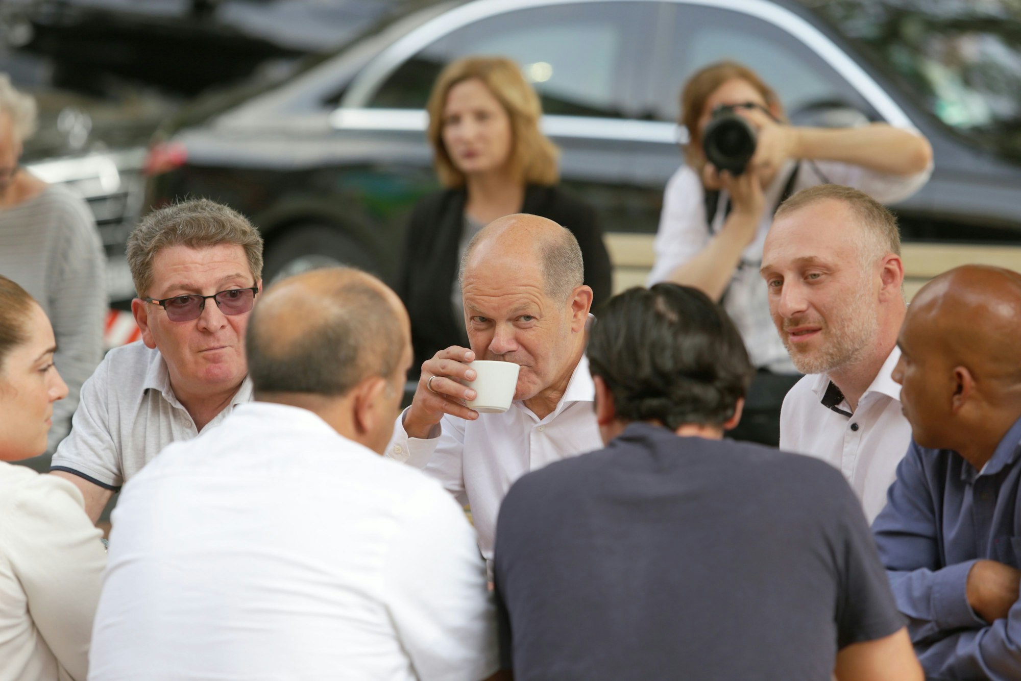 16.08.2023, Düsseldorf: Bundeskanzler Olaf Scholz besucht Büdchen in Düsseldorf Bilk anlässlich des Büdchentages. Der Bundeskanzler sitzt umringt von Menschen an einem Tisch und trinkt aus einer Tasse.