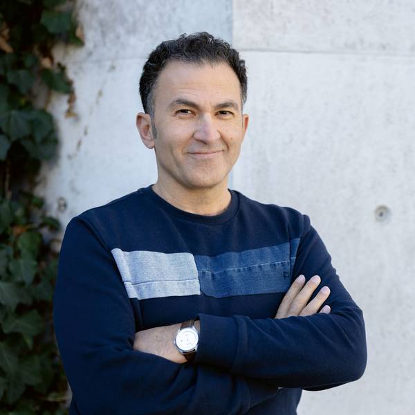 Fatih Çevikkollu steht in einem blauen Pullover vor einer weißen Wand.
