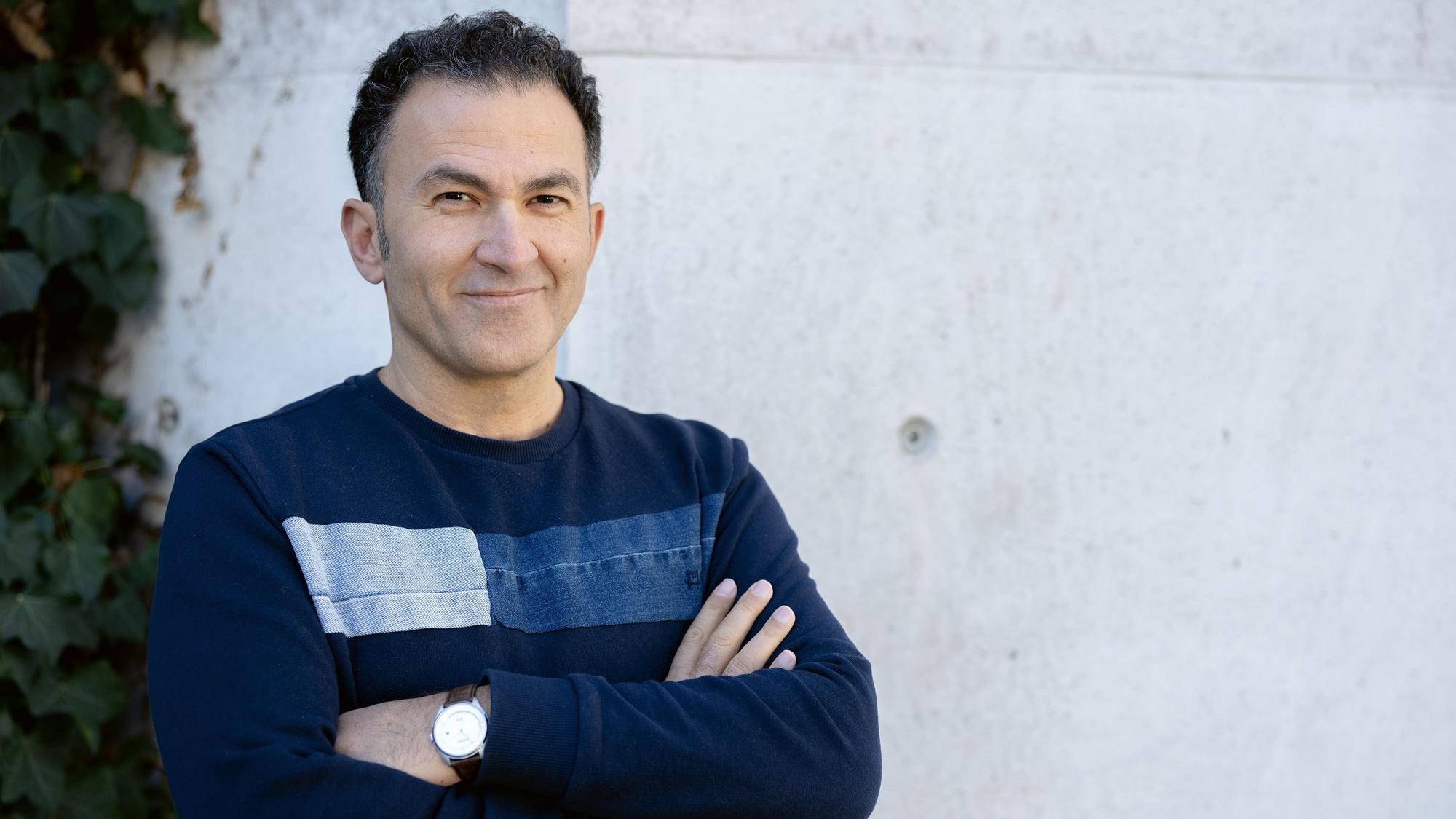 Fatih Çevikkollu steht in einem blauen Pullover vor einer weißen Wand.