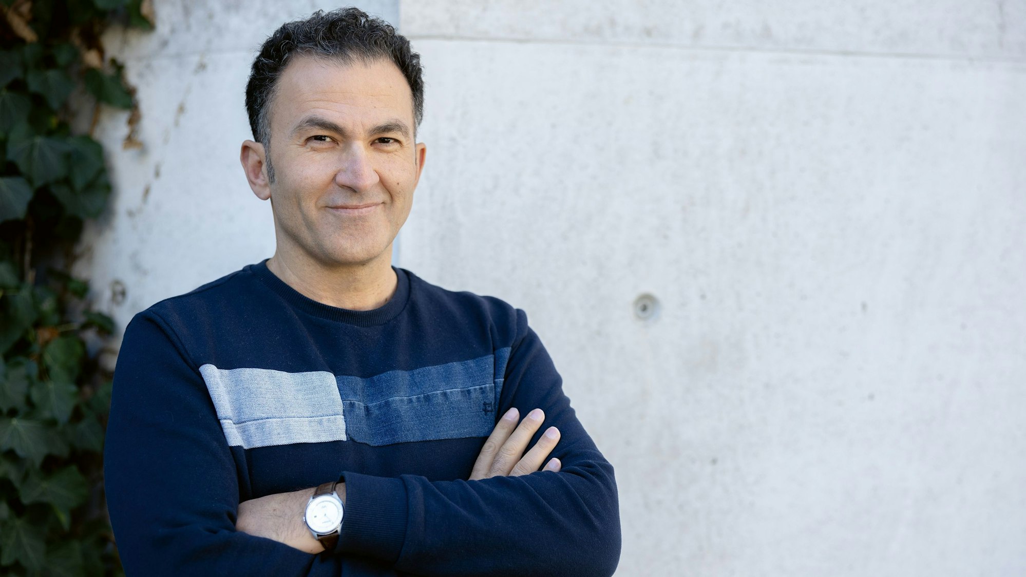 Fatih Çevikkollu steht in einem blauen Pullover vor einer weißen Wand.