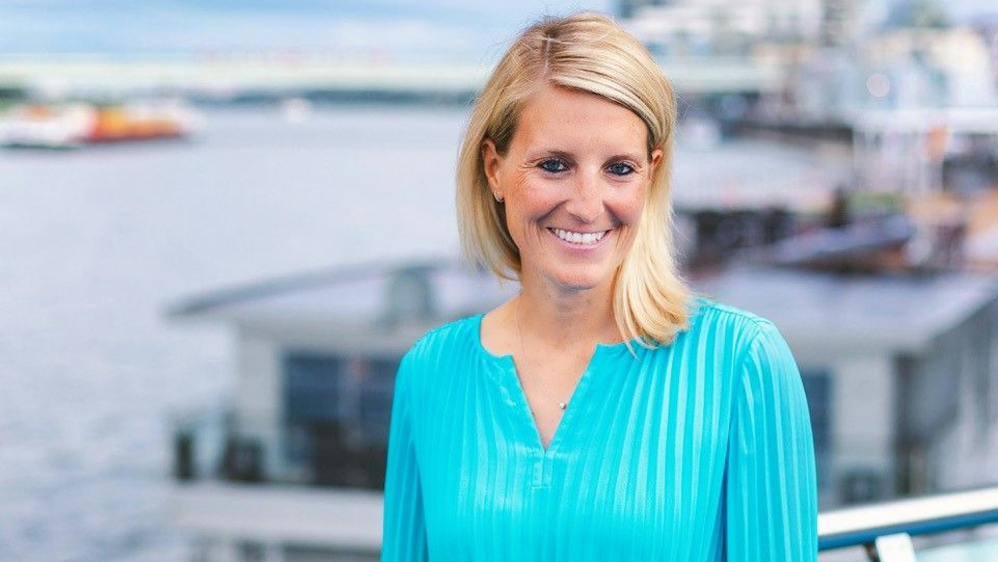 Eine blonde Frau im blauen Kleid steht vor dem Rhein in Köln.