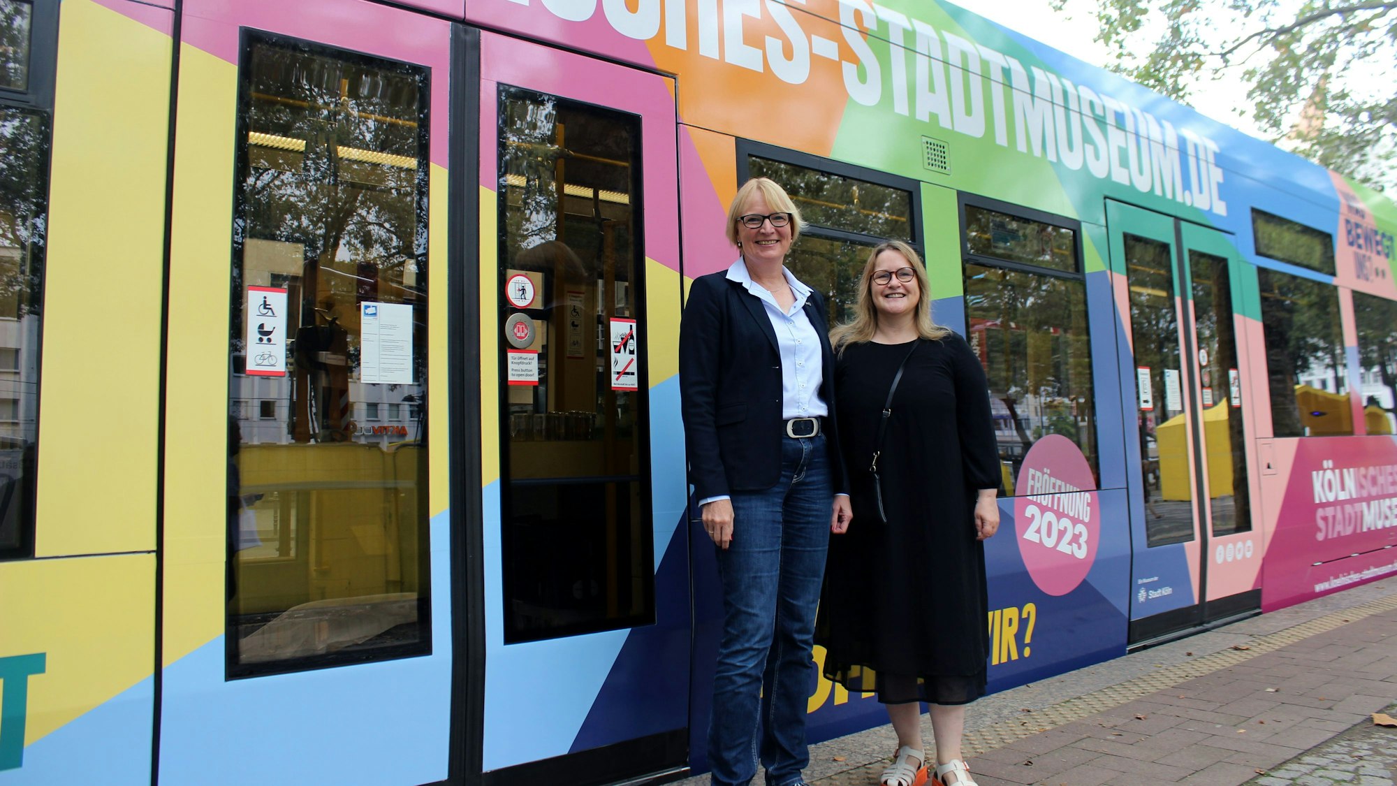 Zwei Frauen, die vor einer bunt bedruckten KVB-Bahn auf dem Neumarkt in Köln stehen.
