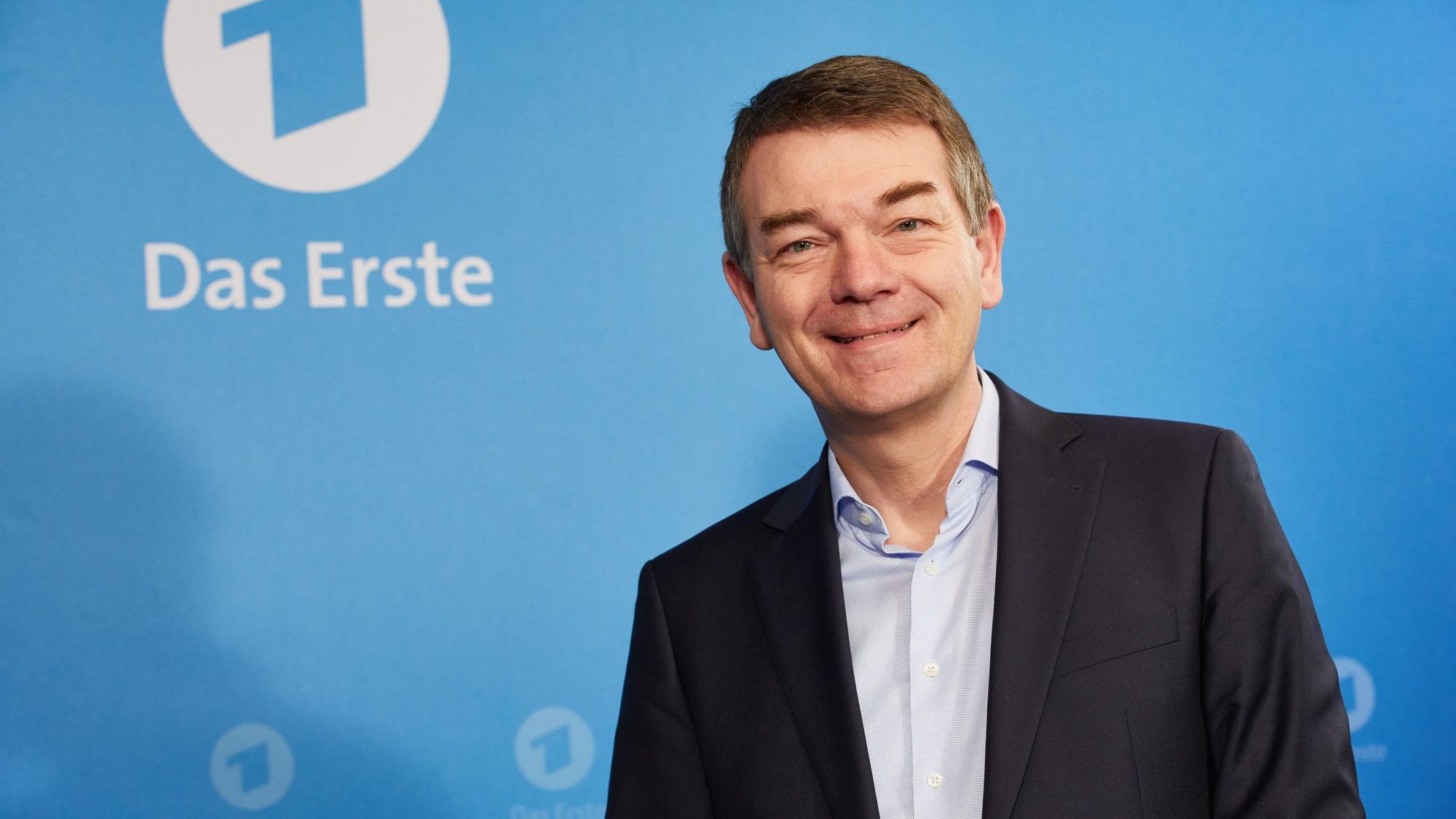 Jörg Schönenborn steht vor einer blauen Wand mit dem Logo des Ersten und lächelt in die Kamera. Er trägt einen dunklen Anzug und ein hellblaues Hemd