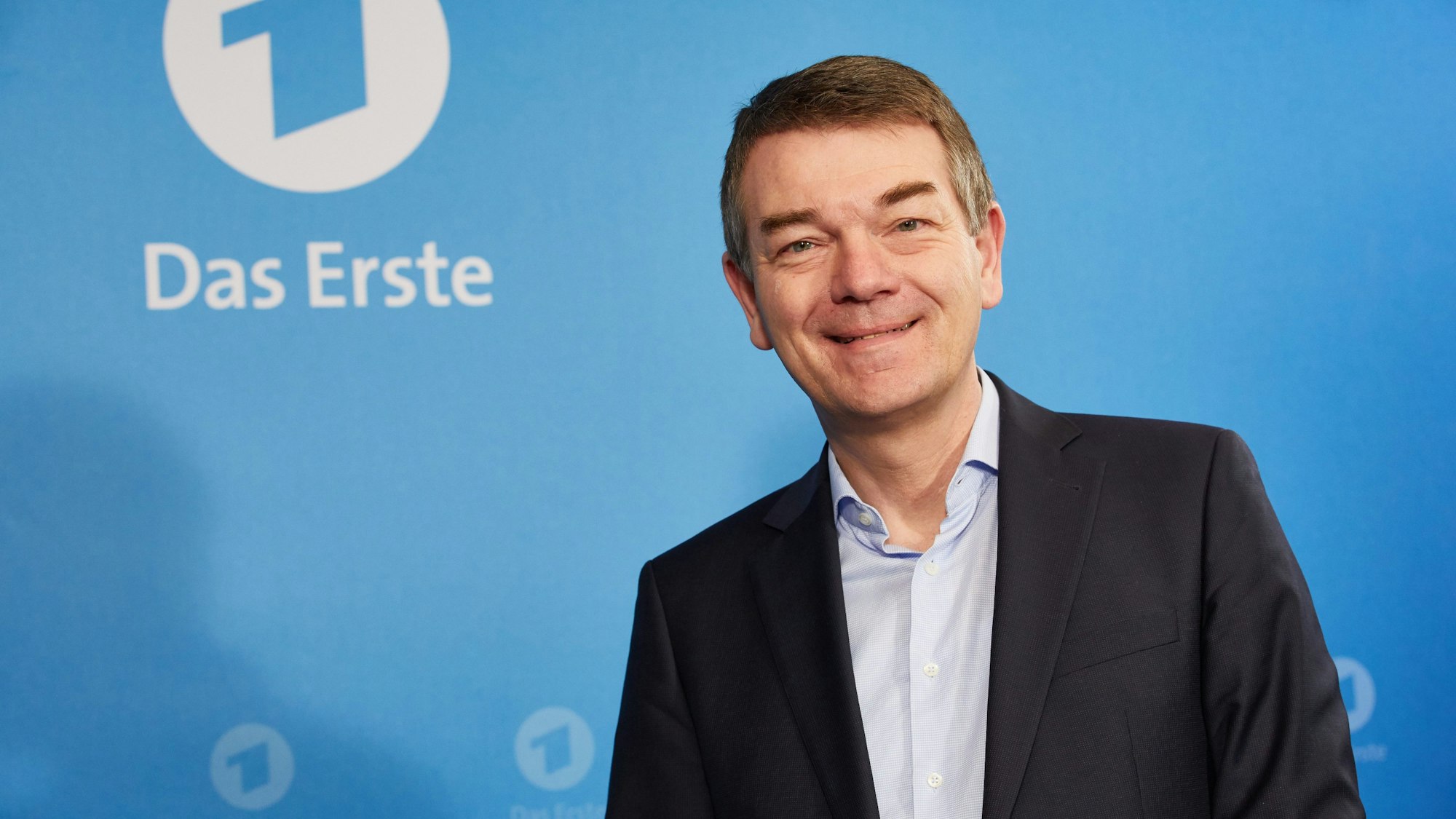 Jörg Schönenborn steht vor einer blauen Wand mit dem Logo des Ersten und lächelt in die Kamera. Er trägt einen dunklen Anzug und ein hellblaues Hemd