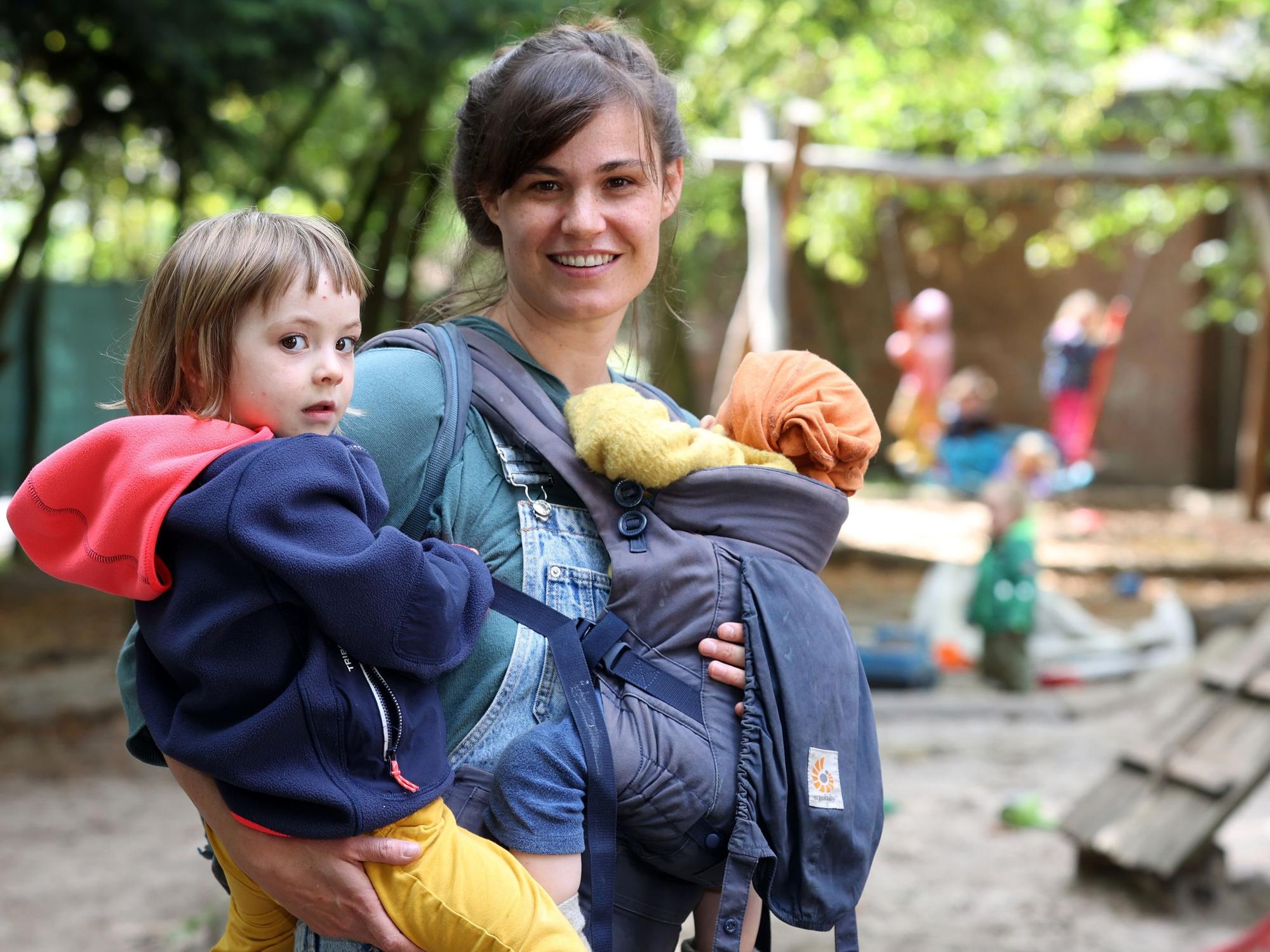 Eine Frau steht mit zwei Kleinkindern auf dem Arm auf dem Außengelände einer Kita.