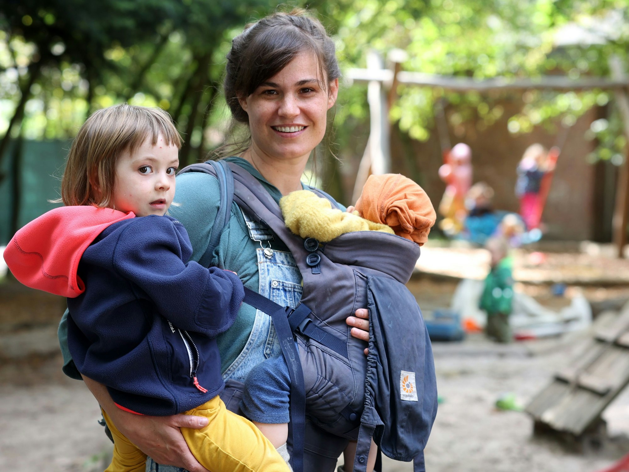 Eine Frau steht mit zwei Kleinkindern auf dem Arm auf dem Außengelände einer Kita.