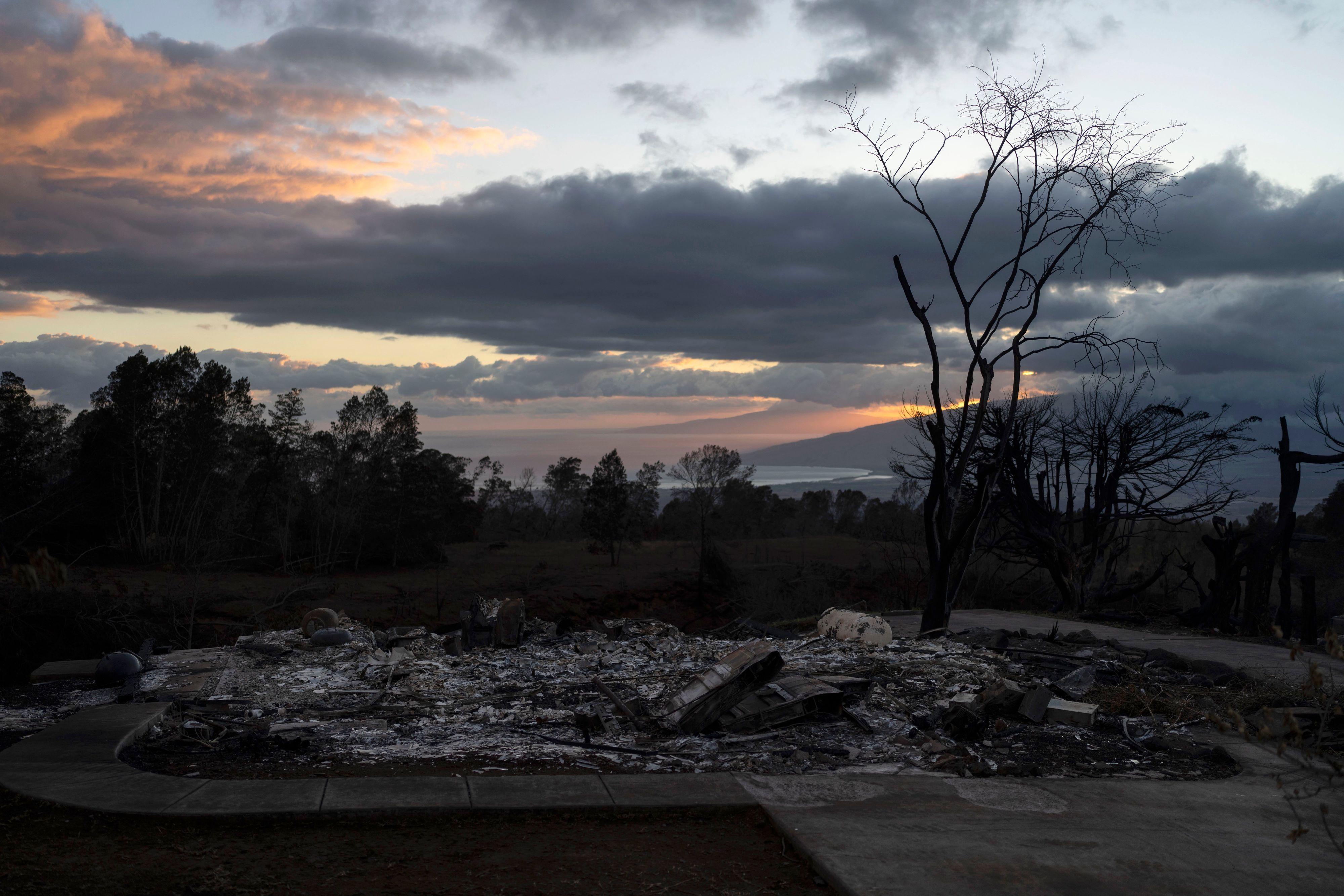 Die abgebrannten Überreste eines Hauses sind im Vordergrund zu sehen, während der Sonnenuntergang den Abendhimmel in Kula rötlich einfärbt.