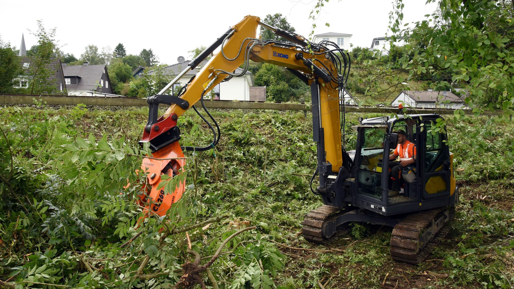 Ein Bagger mit Greifarm steht in einer abgeholzten Böschung und sortiert abgetrennte Äste von Bäumen.