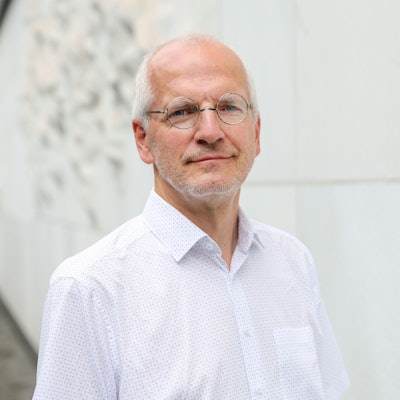 Bönig trägt ein weißes, gepunktetes Hemd und eine runde Brille. Seine Haare sind weiß und er hat einen Dreitagebart.