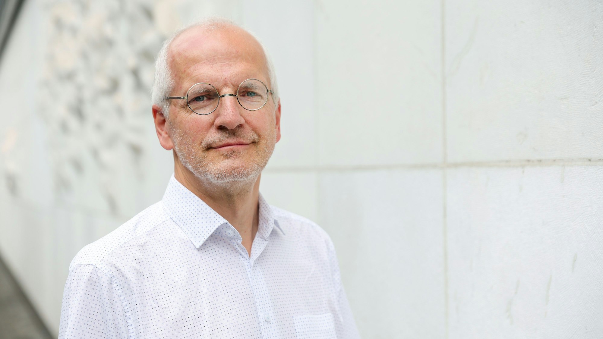 Bönig trägt ein weißes, gepunktetes Hemd und eine runde Brille. Seine Haare sind weiß und er hat einen Dreitagebart.