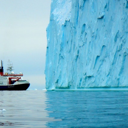 Ein Forschungsschiff fährt in der Antarktis vor einem großen blauen Eisberg entlang. Im Hintergrund sind weitere Eisschollen zu sehen. (Symbolbild)