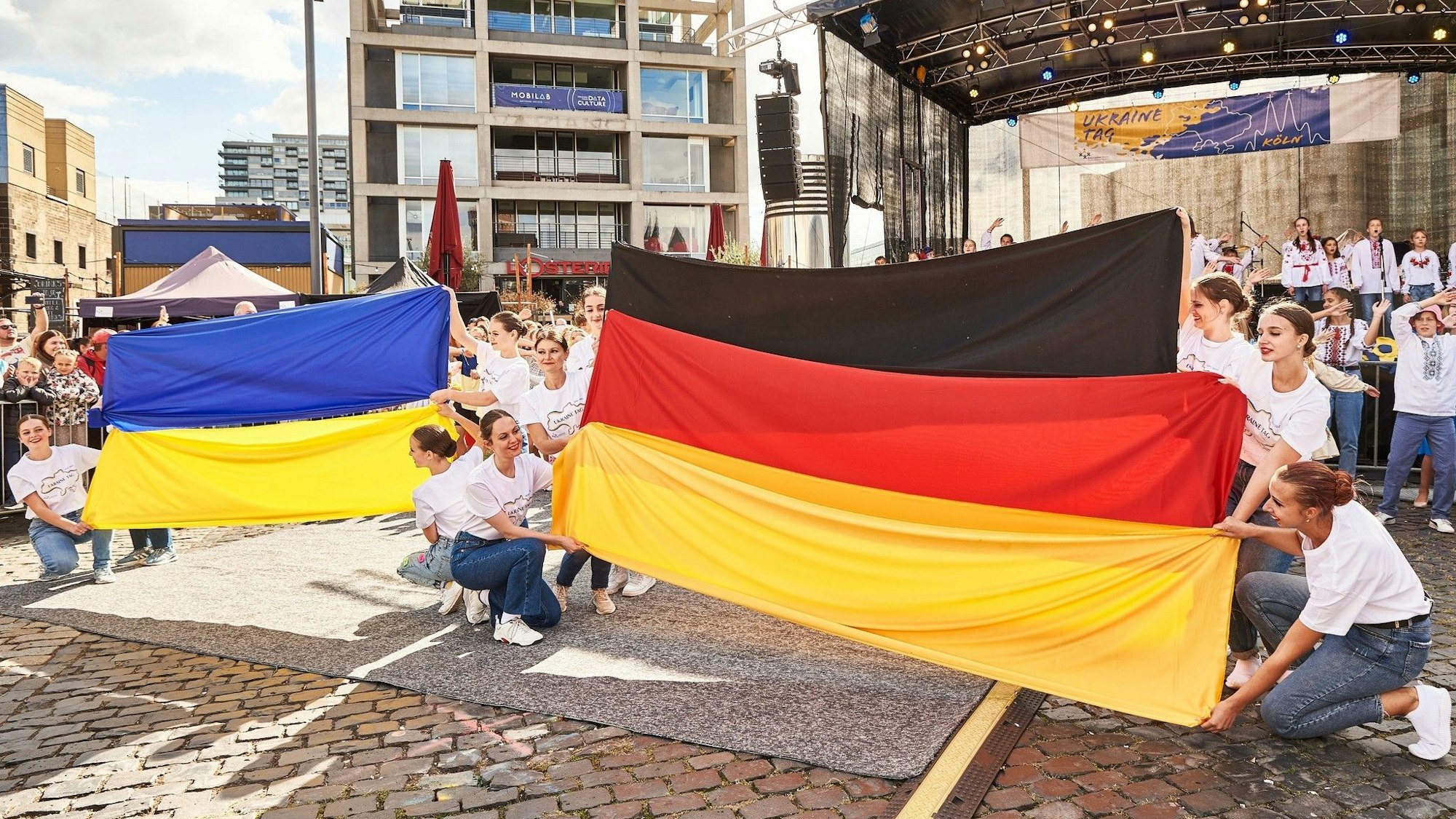 Junge Frauen halten beim Ukraine Tag 2022 vor einer Bühne am Schokoladenmuseum die ukrainische und die deutsche Nationalfahne in den Händen.