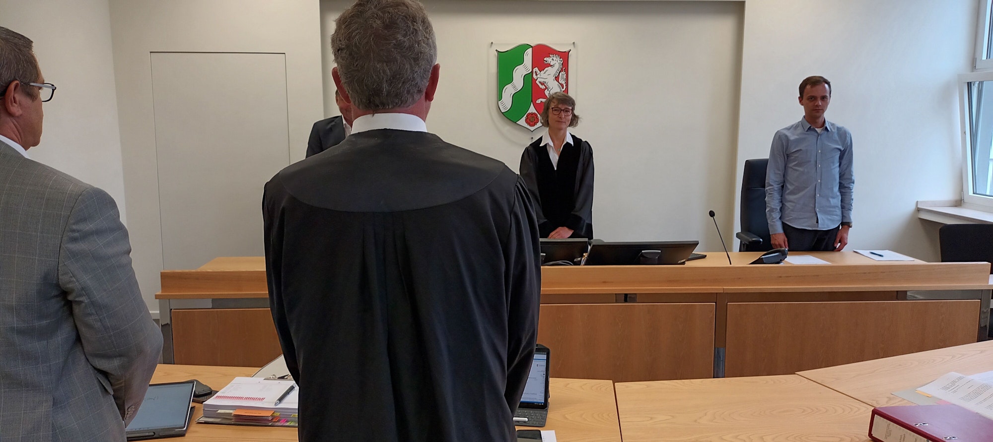 In einem Gerichtssaal stehen eine Richterin und zwei ehrenamtliche Richter sowie die Beklagten-Vertreter