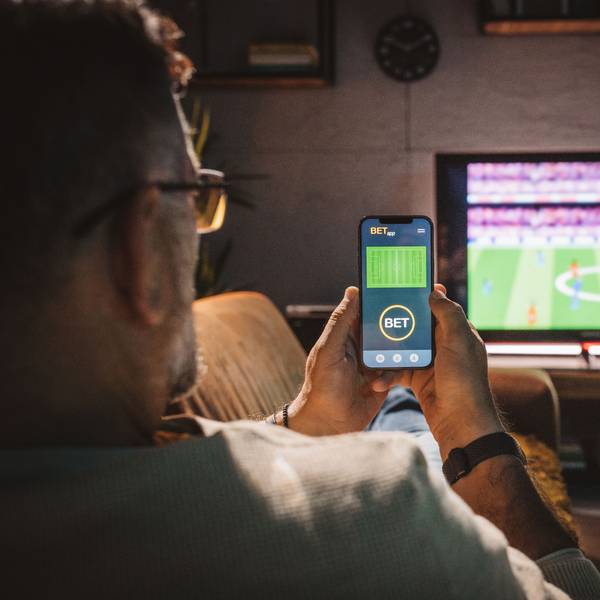 Ein Mann sitzt auf einer Couch, guckt ein Fußballspiel und hält ein Handy in den Händen, in dem er eine Sportwette platziert.