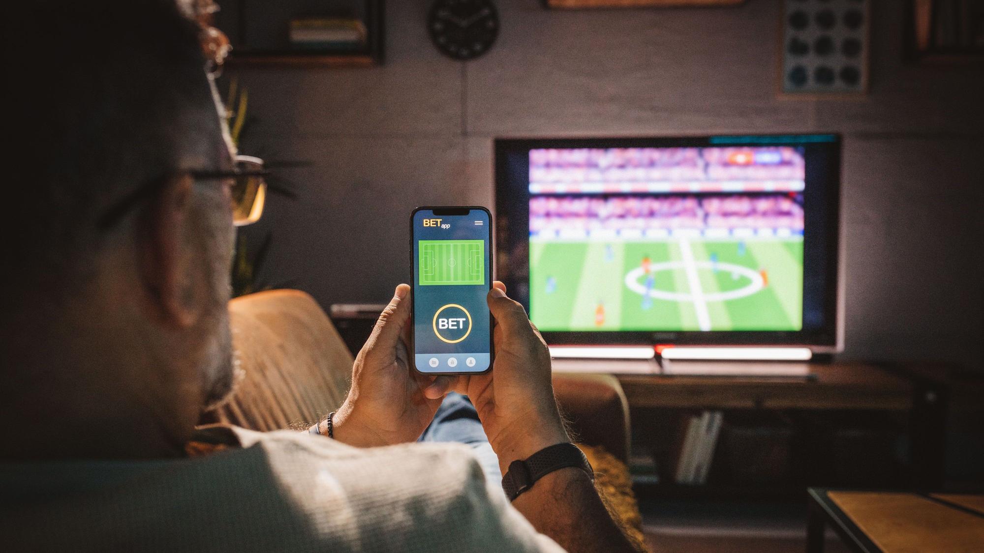 Ein Mann sitzt auf einer Couch, guckt ein Fußballspiel und hält ein Handy in den Händen, in dem er eine Sportwette platziert.
