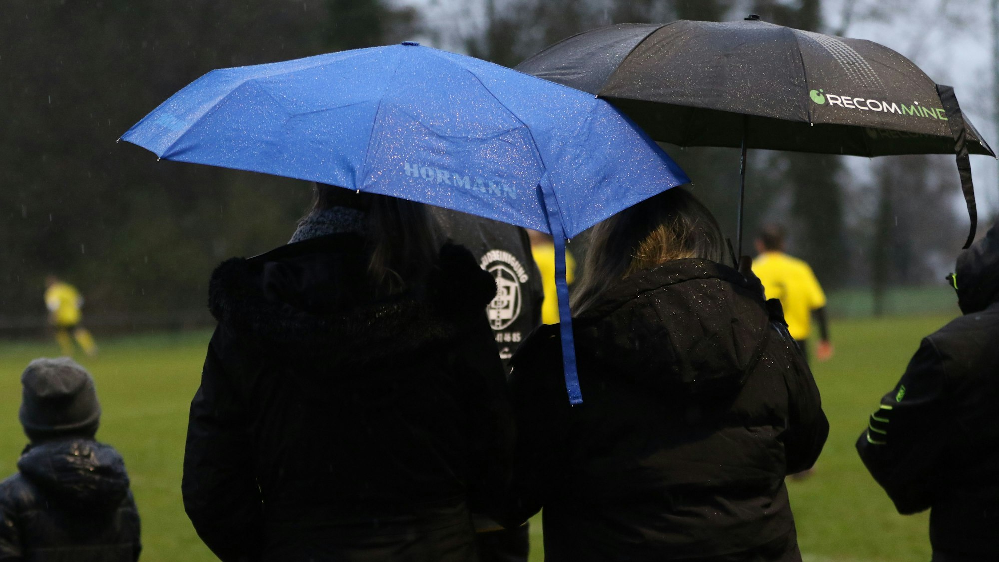 Zwei Zuschauerinnen stehen mit Regenschirm am Rande eines Fußballspiels.