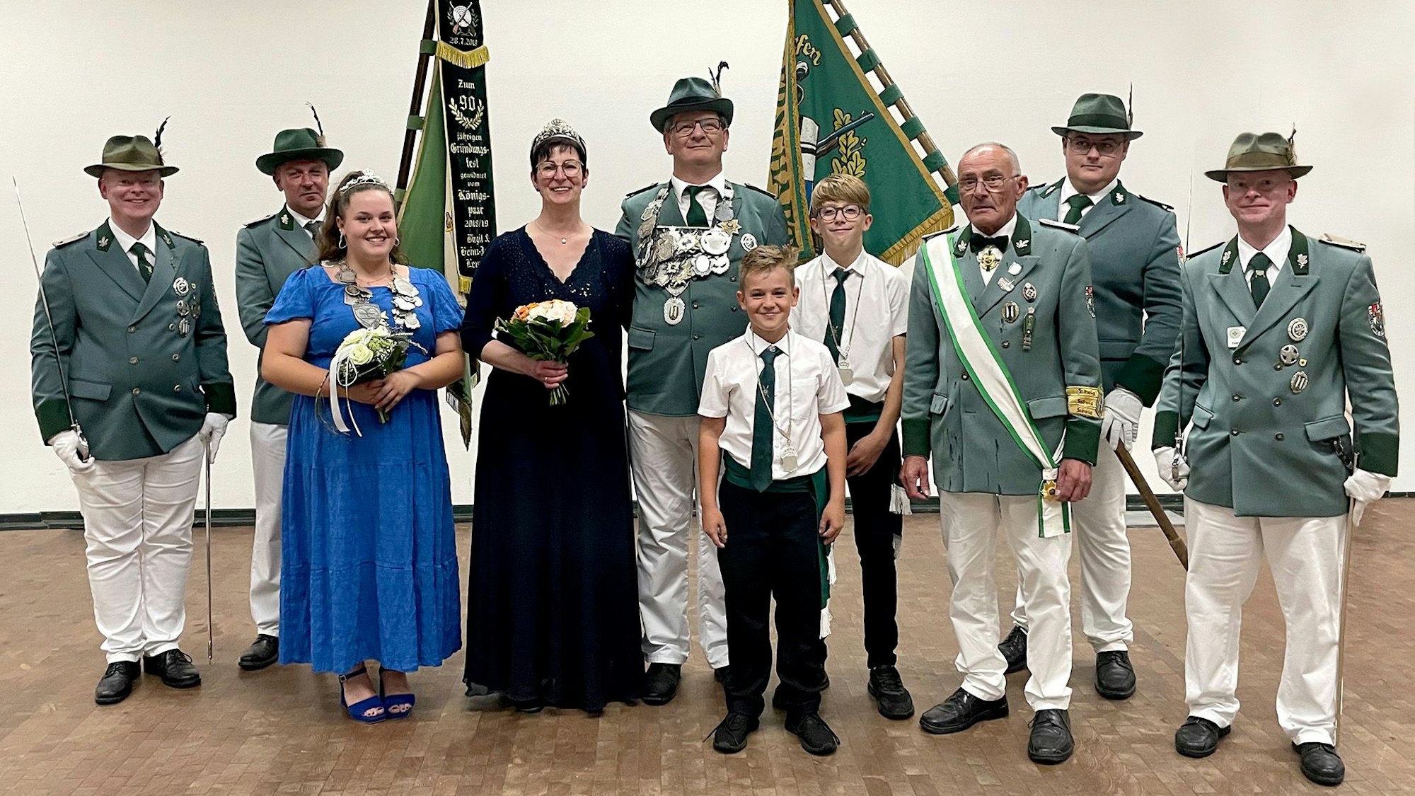 Zehn Mitglieder eines Schützenvereins stehen nebeneinander. Die Damen tragen Kleider, die Herren grüne Uniformen. Zwei Männer tragen grüne Fahnen.