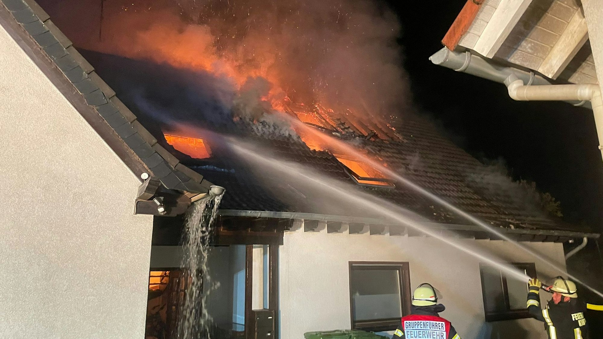 Feuerwehrleute löschen mit Wasser die Flammen, die aus dem Dach eines Hauses schlagen.