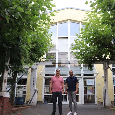 Zwei Männer stehen vor dem Eingang eines großen gelben Gebäudes mit verglasten Eingangstüren und vielen Fenstern.