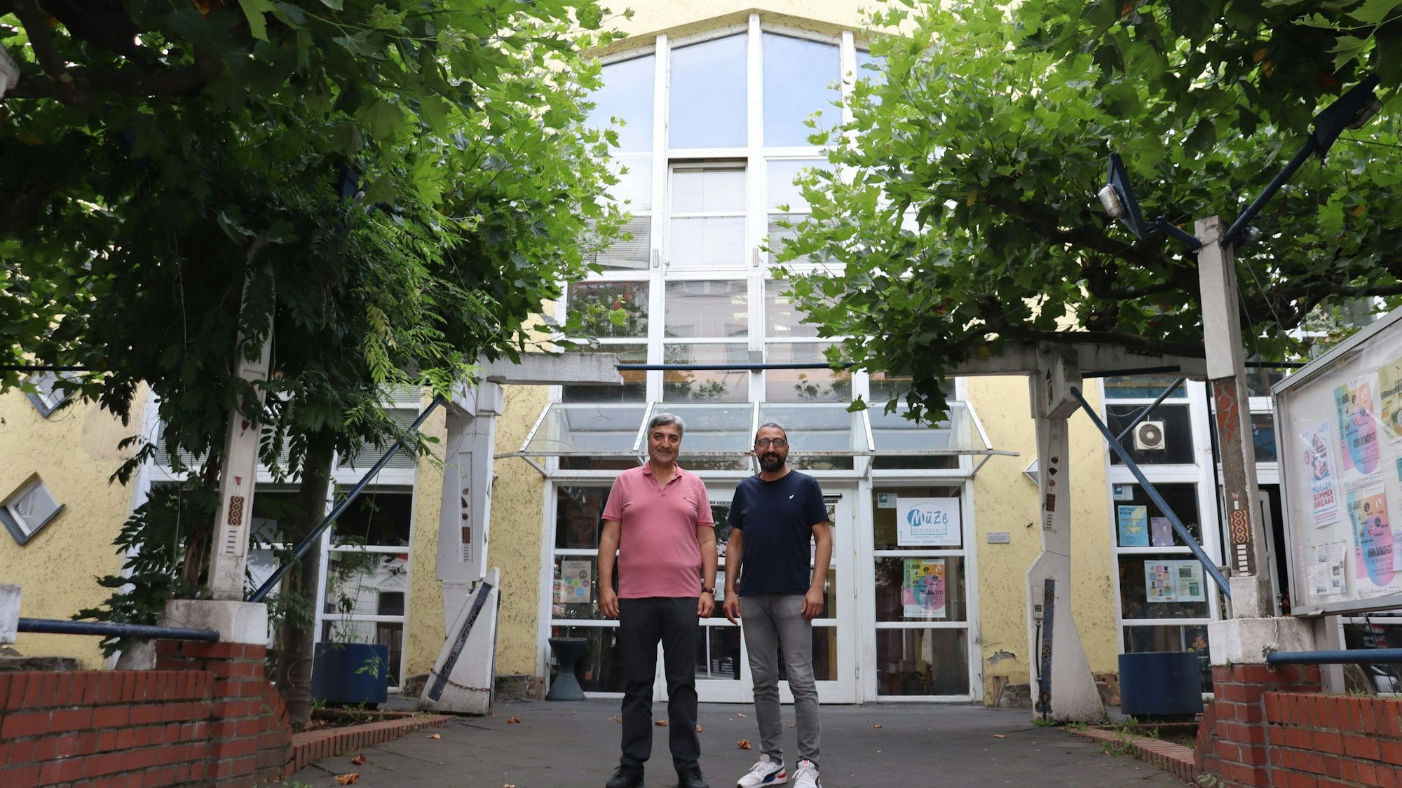 Zwei Männer stehen vor dem Eingang eines großen gelben Gebäudes mit verglasten Eingangstüren und vielen Fenstern.