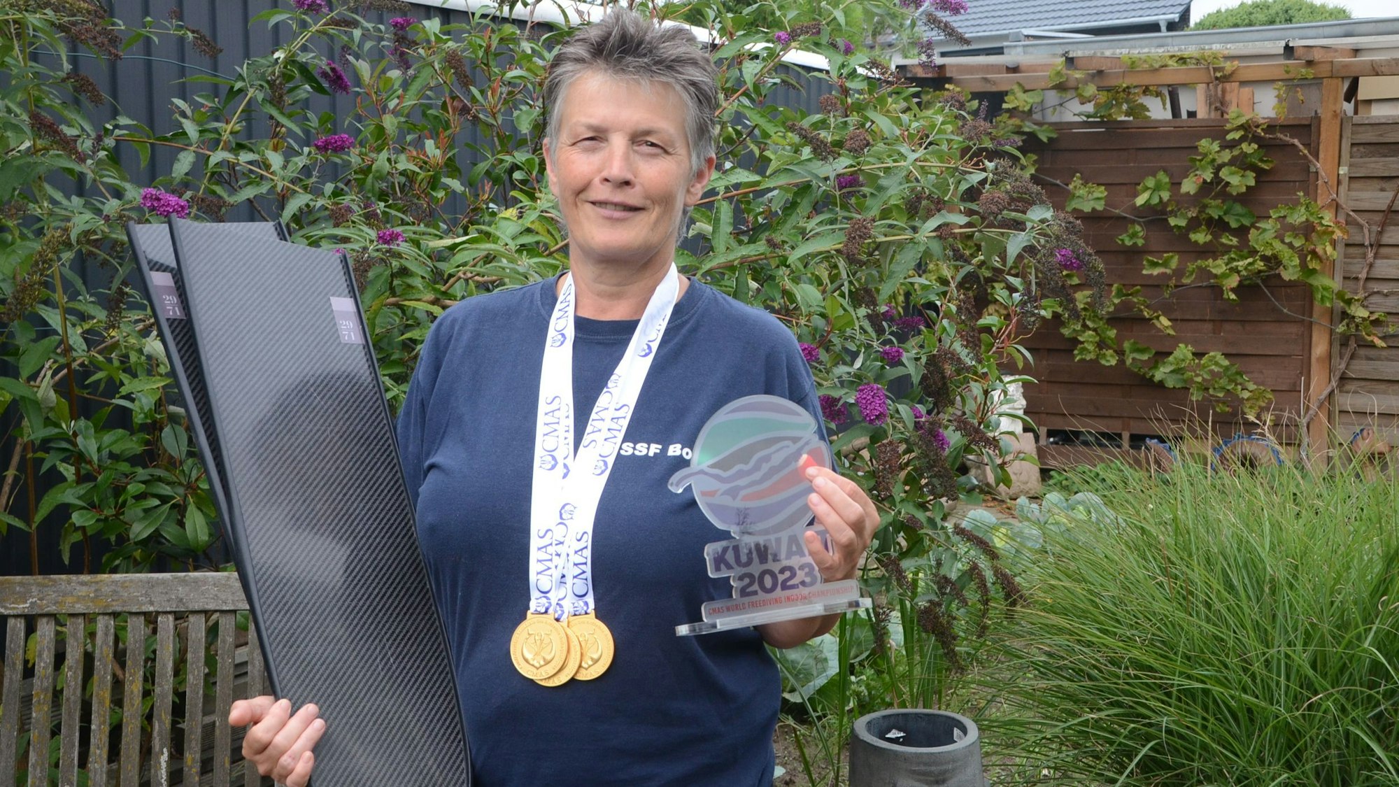 Eine Frau mit kurzen grauen Haaren steht in ihrem Garten. Sie hat drei Goldmedaillen um den Hals hängen; im rechten Arm trägt sie Schwimmflossen. In der linken Hand hält sie eine bunte Trophäe von der Weltmeisterschaft