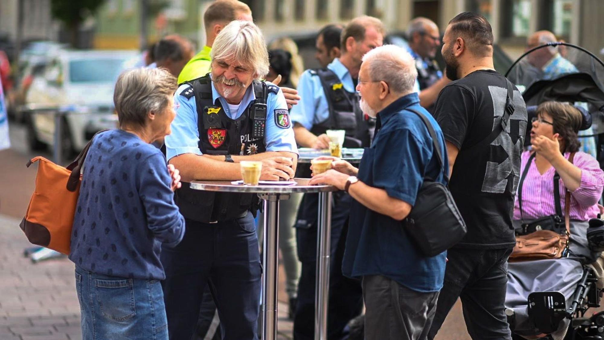 Der Bezirksdienstbeamte Roger Kath steht mit zwei Euskirchener Bürgern an einem Stehtisch. Sie haben jeweils ein Glas mit Kaffee vor sich stehen.