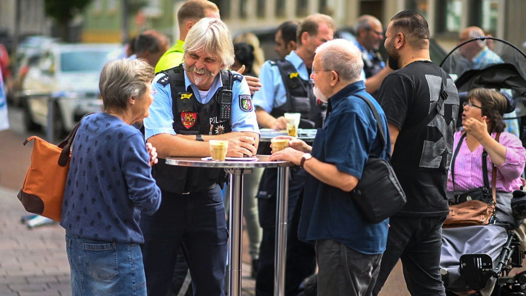Der Bezirksdienstbeamte Roger Kath steht mit zwei Euskirchener Bürgern an einem Stehtisch. Sie haben jeweils ein Glas mit Kaffee vor sich stehen.