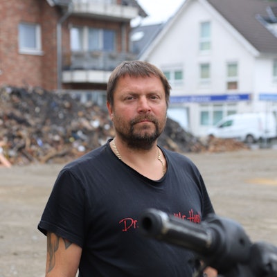 Ein bärtiger Mann im schwarzen T-Shirt steht vor einem großen Schutthaufen.