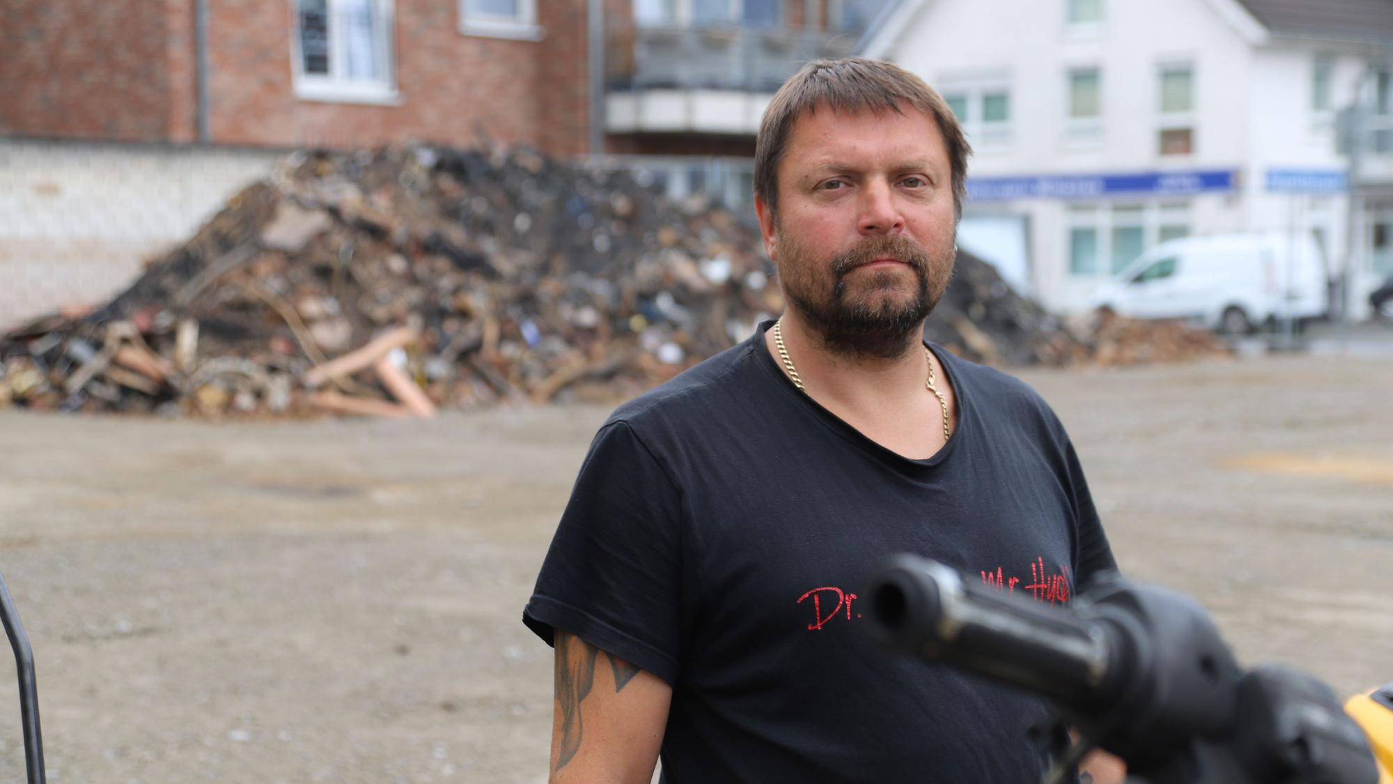 Ein bärtiger Mann im schwarzen T-Shirt steht vor einem großen Schutthaufen.