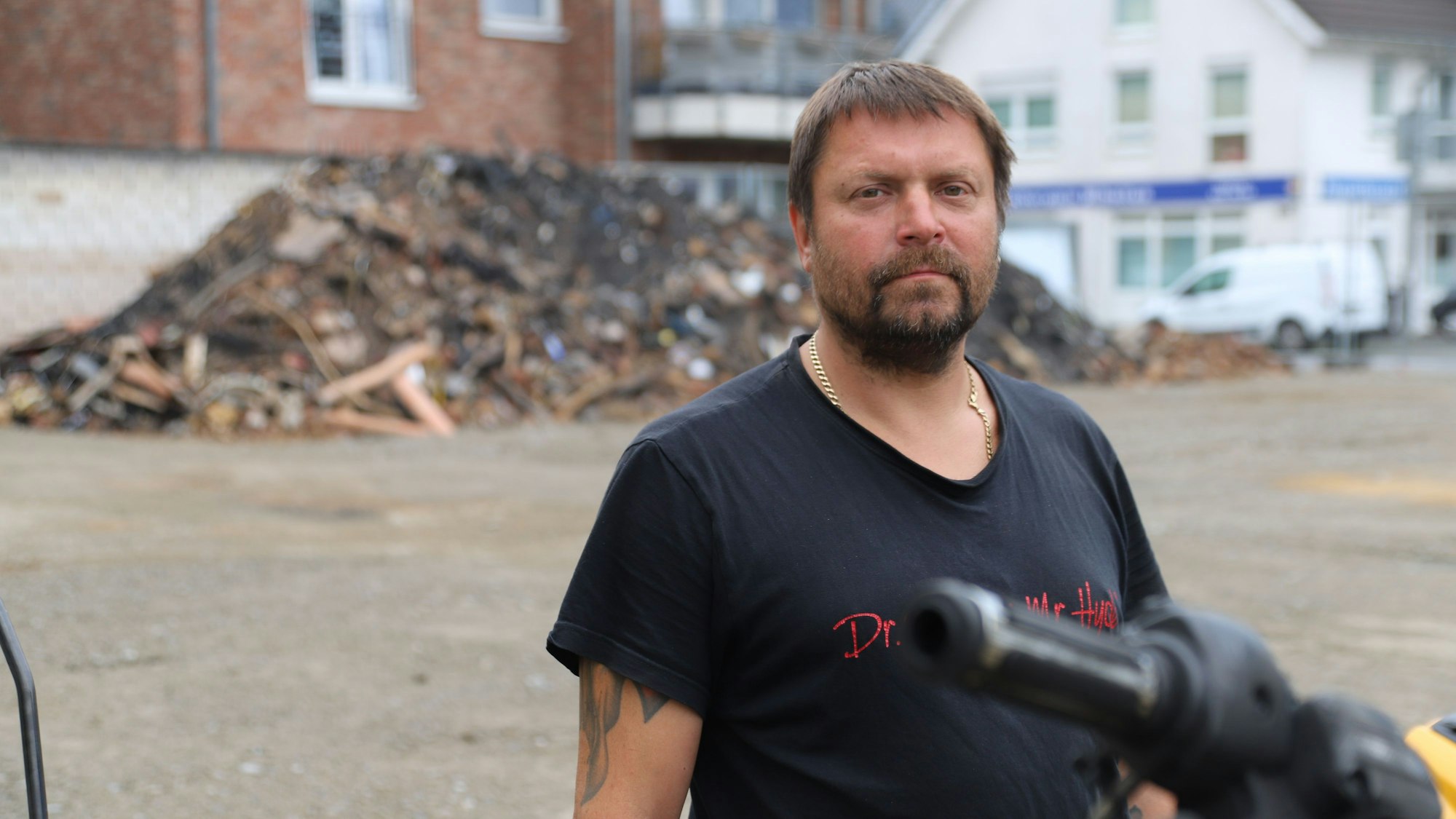 Ein bärtiger Mann im schwarzen T-Shirt steht vor einem großen Schutthaufen.