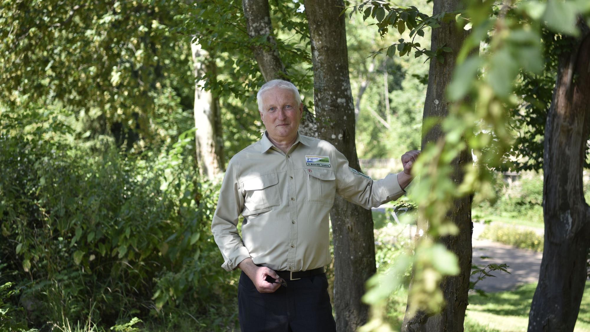 Dr. Michael Röös steht in einer Baumgruppe und stützt sich mit der Hand an einem Stamm ab. Er trägt eine dunkle Hose und ein beiges Hemd mit dem Logo des Nationalpark Eifel auf der Brust.