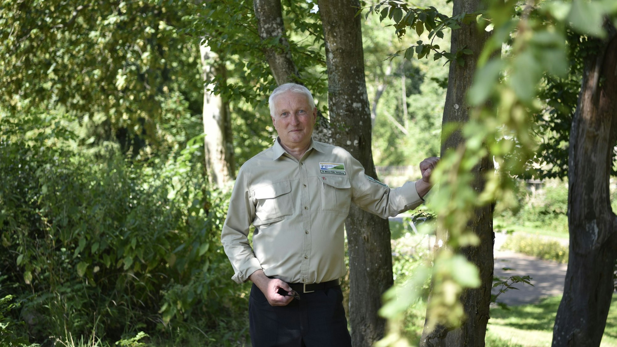 Dr. Michael Röös steht in einer Baumgruppe und stützt sich mit der Hand an einem Stamm ab. Er trägt eine dunkle Hose und ein beiges Hemd mit dem Logo des Nationalpark Eifel auf der Brust.