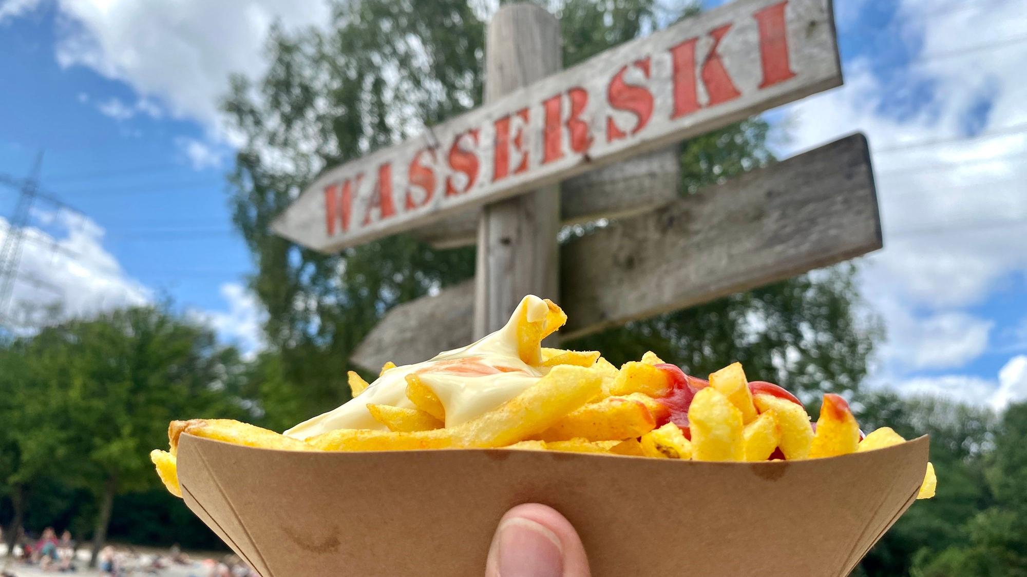 Pommes Rot-Weiß liegen in einer offenen Pappschachtel, im Hintergrund weist ein Holzschild den Weg zum Wasserski.
