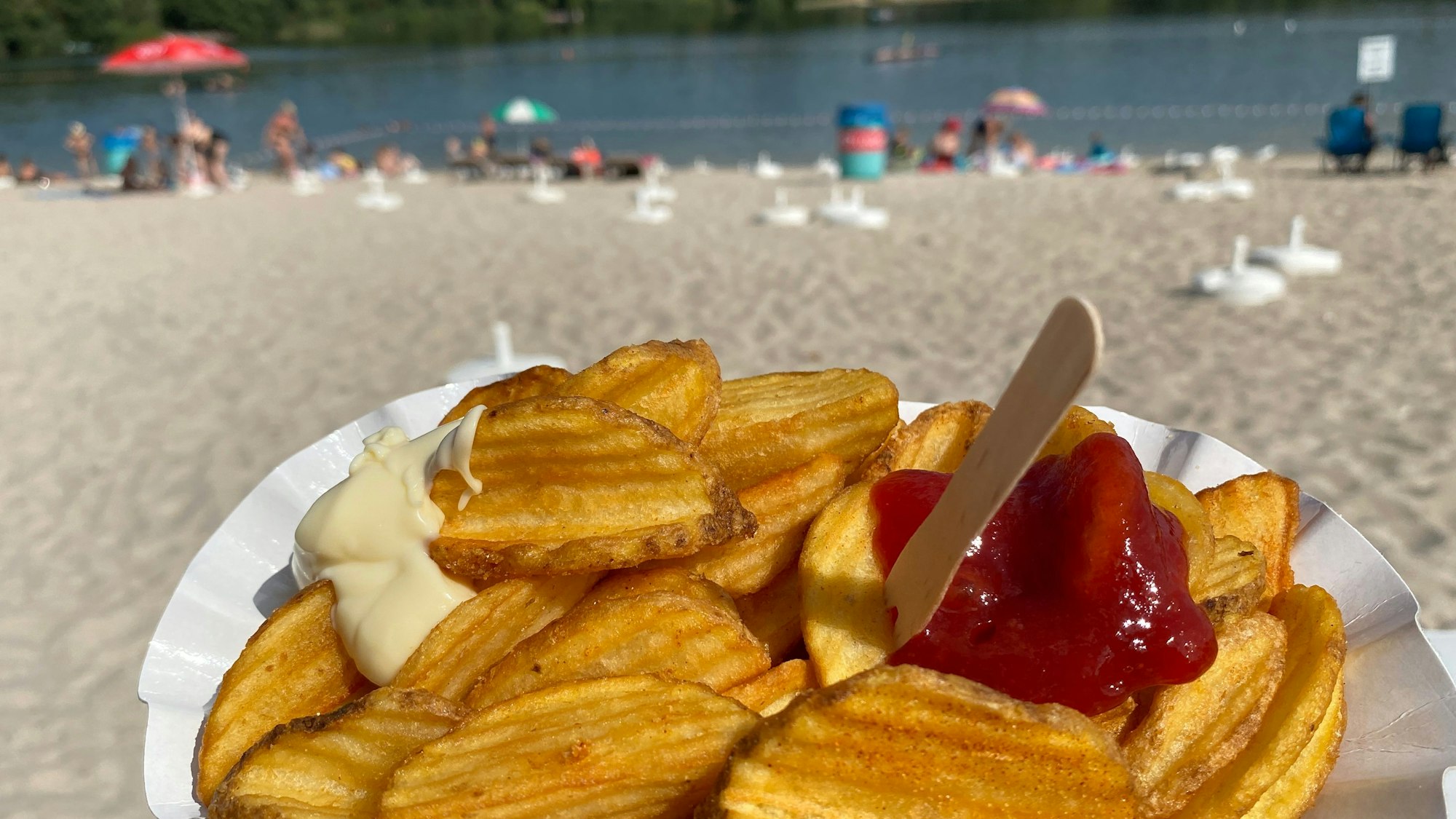 Auf einer Pappschale liegen frittierte geriffelte Kartoffeln mit Mayo und Ketcheup, im Hintergrund ist ein Sandstrand und ein See zu sehen.