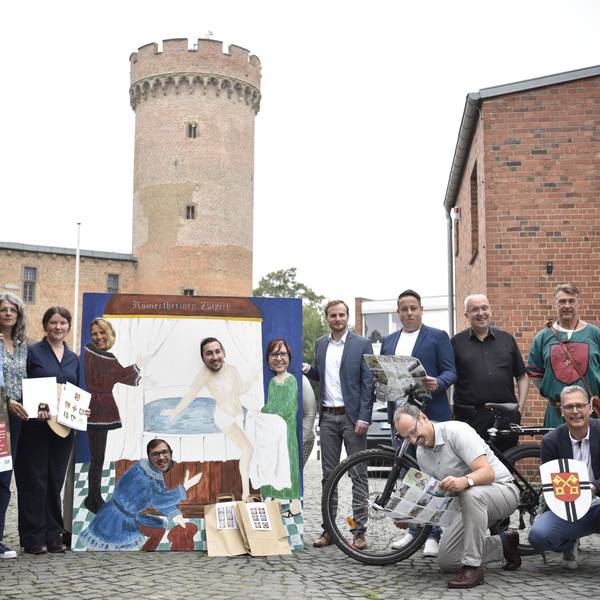Eine Gruppe von Menschen steht auf einem Platz. Im Hintergrund ist ein mittelalterlicher Turm zu erkennen.