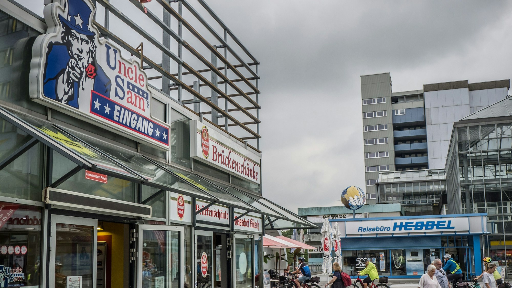 Der Eingangsbereich der Sportsbar Uncle Sam's am Rialto Boulevard ist neben der Brückenschänke zu sehen. Davor laufen Menschen, im Hintergrund ist das Reisebüro Hebbel.
