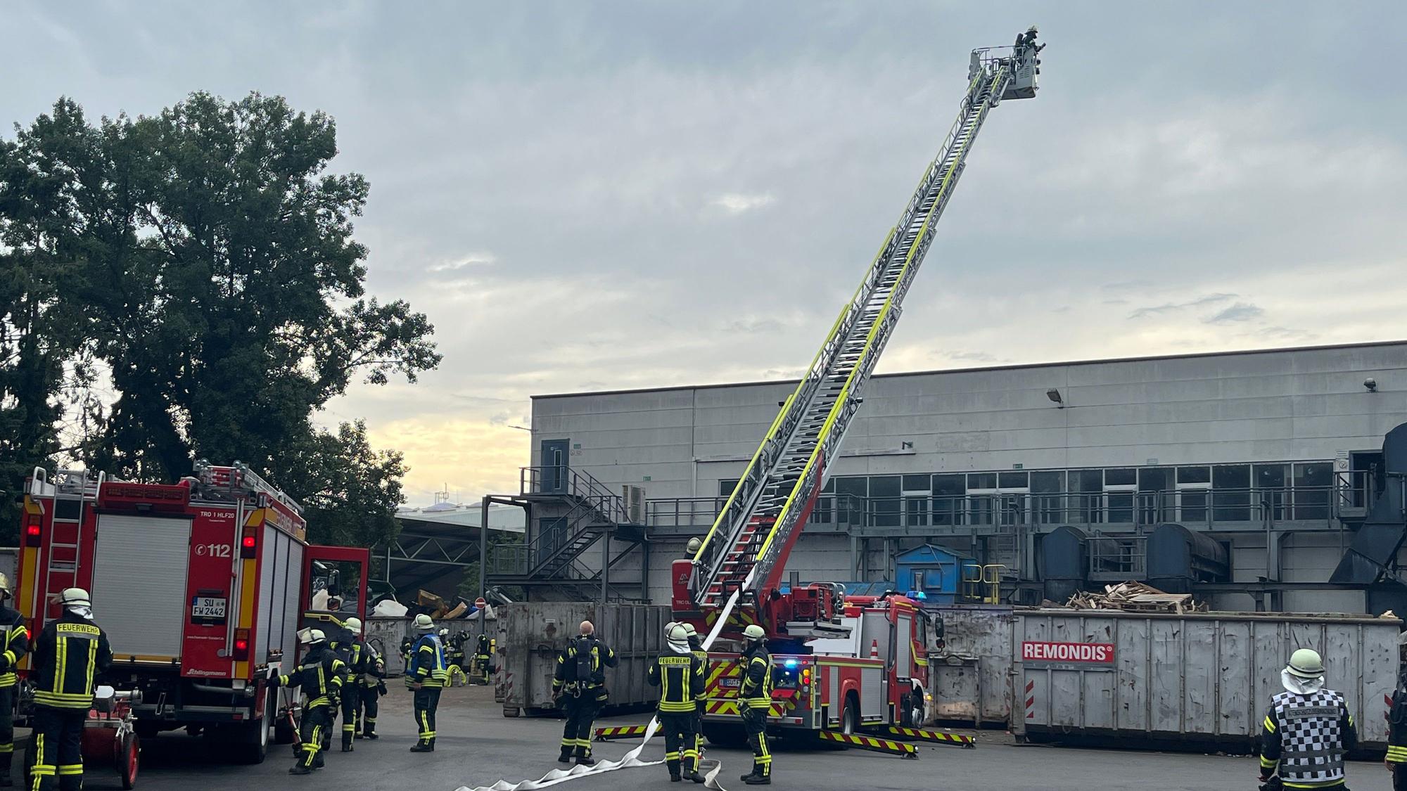 Feuerwehrleute stehen mit mehreren Löschfahrzeugen und einer Drehleiter an einer Industriehalle, Container mit dem Schriftzug Remondis stehen davor.