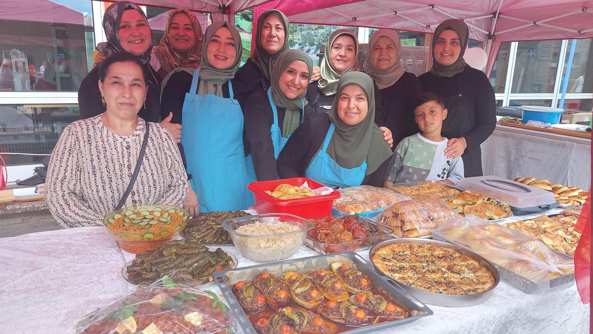Lächelnde Frauen, fast alle mit Kopftüchern, stehen hinter einem großen Tisch, auf dem Platten und Schüsseln voller Essen stehen.