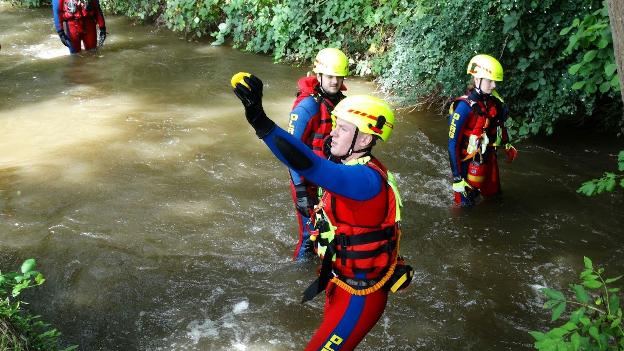 Ein Mann in rot-blauem Neoprenanzug und mit Helm steht im Wasser und hebt eine Gummiente in die Höhe.