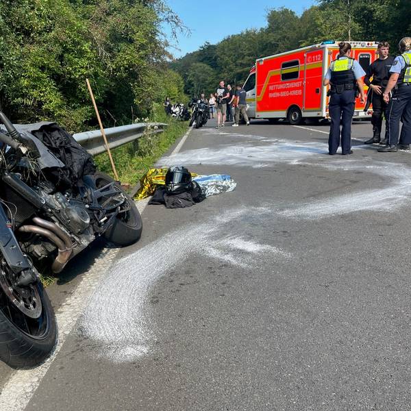 Ein beschädigtes Motorrad lehnt an der Leitplanke einer Straße, weiße Spuren sind auf dem Asphalt. Motorradkleidung und ein Helm liegen auf der Straße. Polizisten und ein Rettungswagen stehen im Hintergrund.