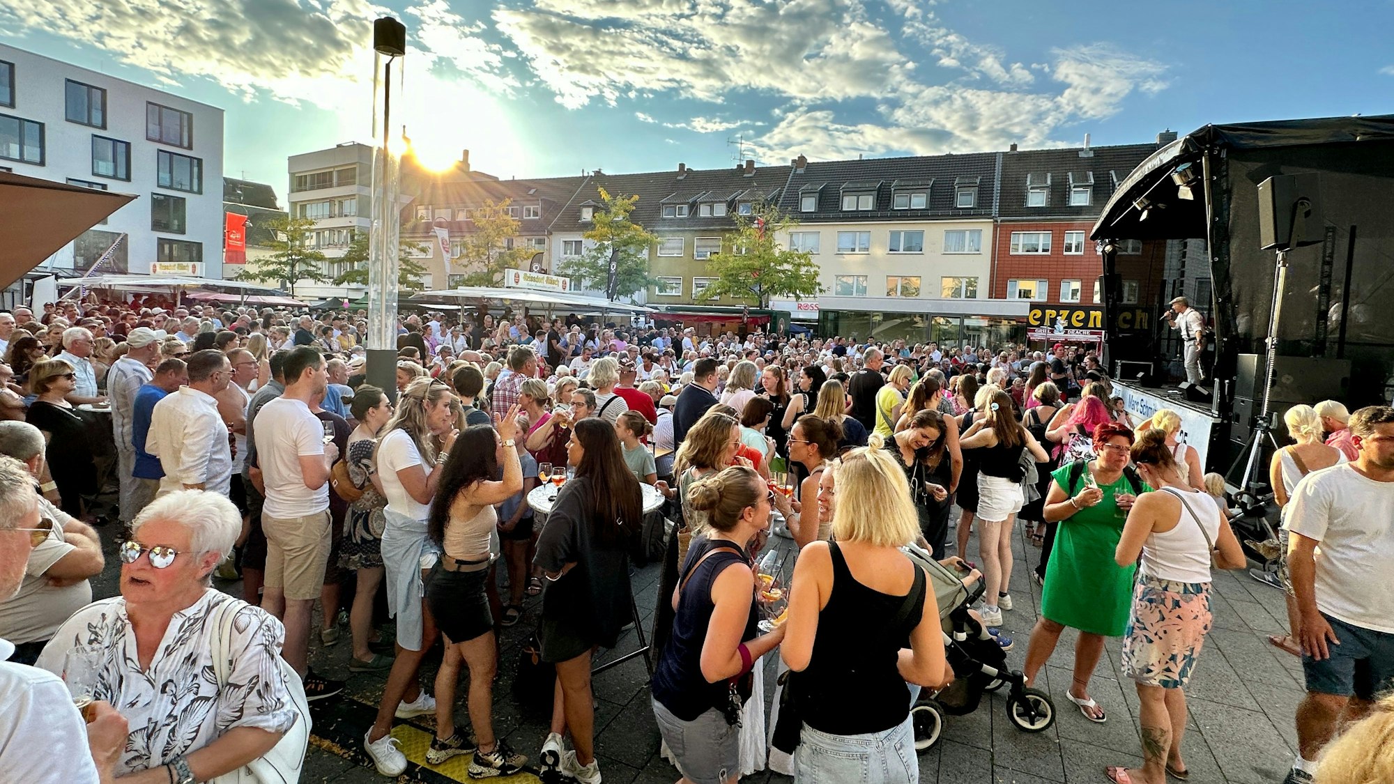 Blick auf den sonnigen Maternusplatz, der mit Menschen gefüllt ist. Sie stehen vor einer Bühne, auf der die Klüngelköpp auftreten.