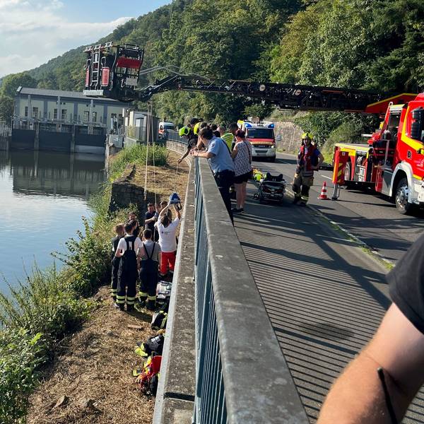 An einem breiten Kanal stehen mehrere Rettungskräfte an einer Mauer, die zu einer höher gelegenen Straße gehört. Dort steht die ausgefahrene Drehleiter der Feuerwehr.