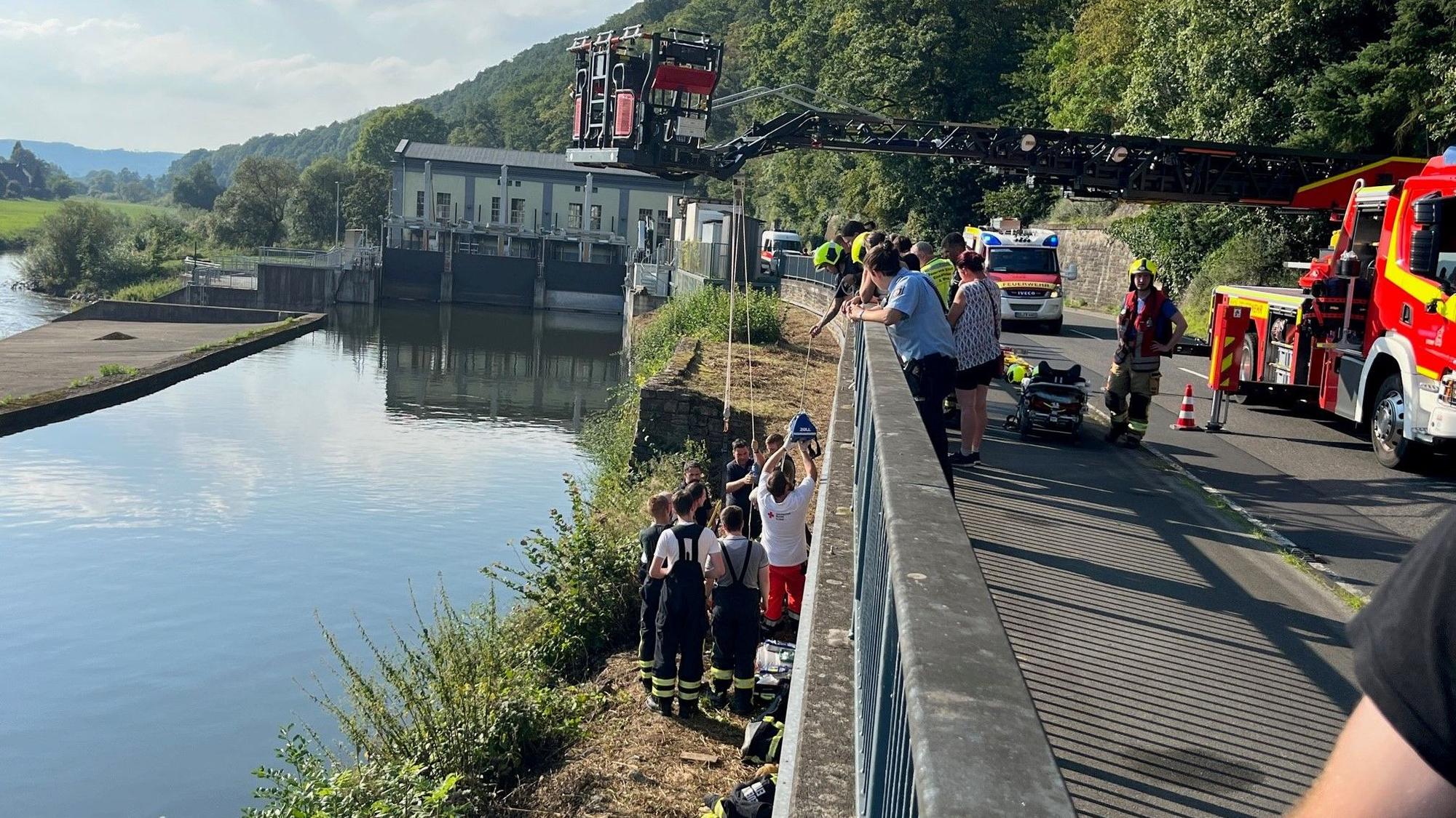 An einem breiten Kanal stehen mehrere Rettungskräfte an einer Mauer, die zu einer höher gelegenen Straße gehört. Dort steht die ausgefahrene Drehleiter der Feuerwehr.