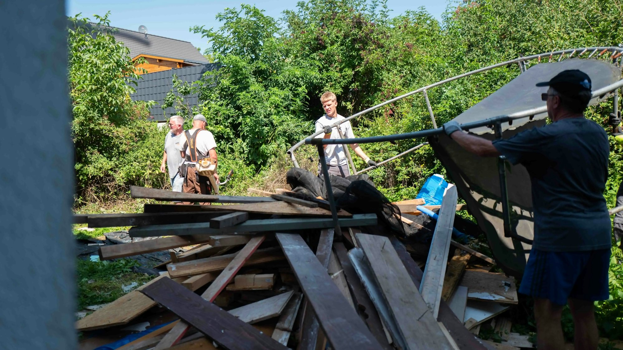 Im Garten tragen zwei Helfer das Gerüst eines Trampolins.