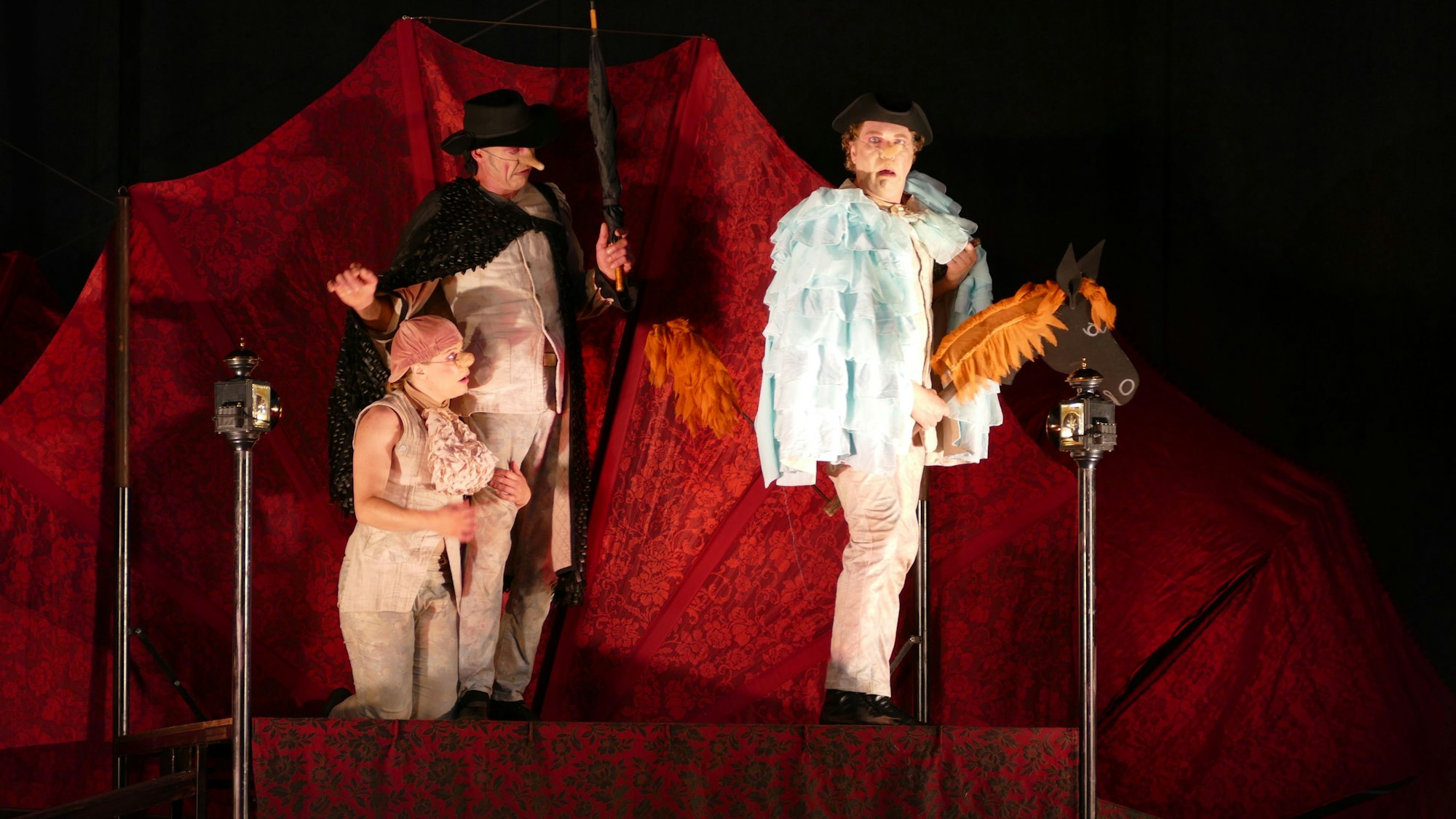 Vor einem roten Hintergrund stehen drei Schauspieler mit Steckenpferd und Regenschirm auf der Bühne und schneiden Grimassen.